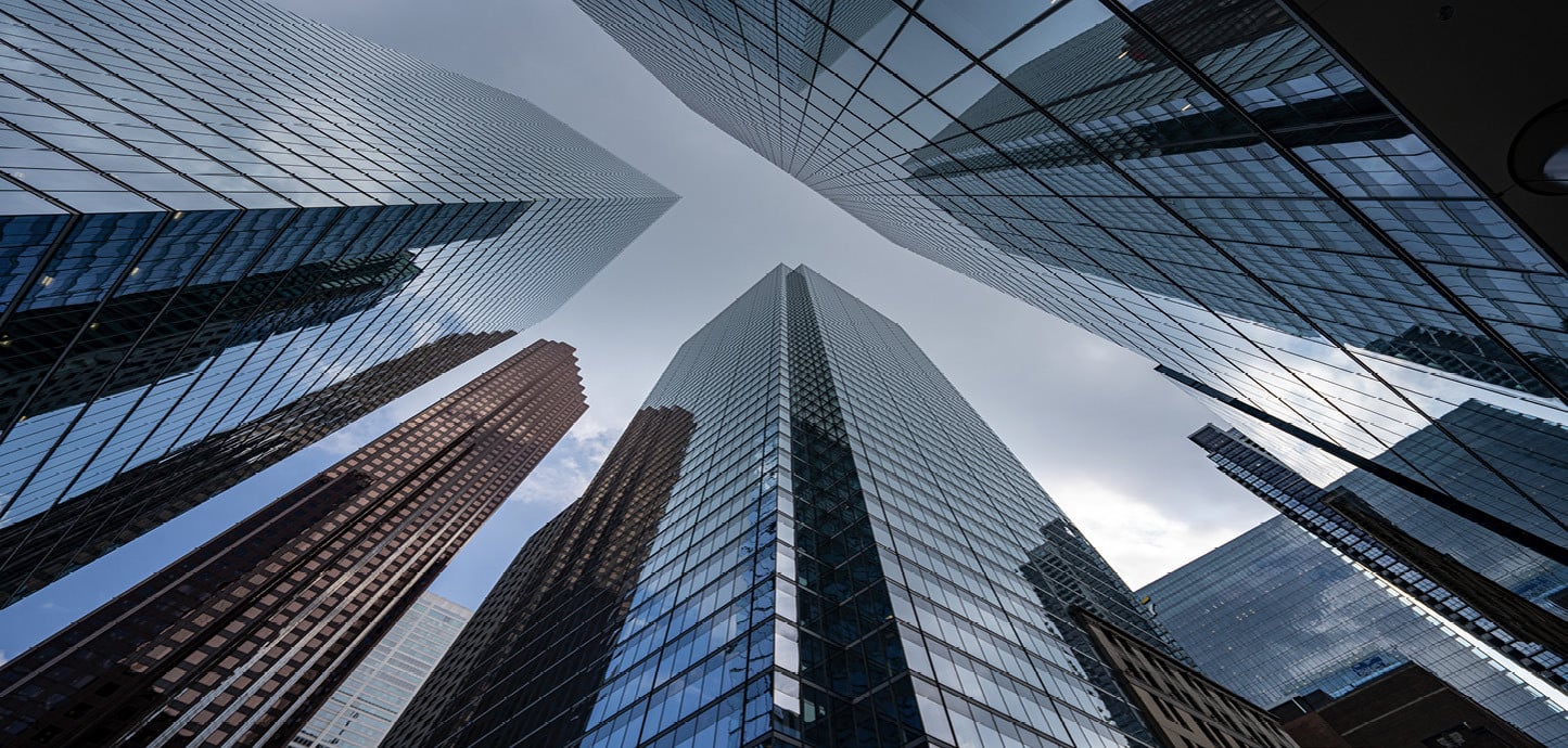 Tall glass and steel office buildings photographed from below, with the façades stretching upwards towards a bright sky.